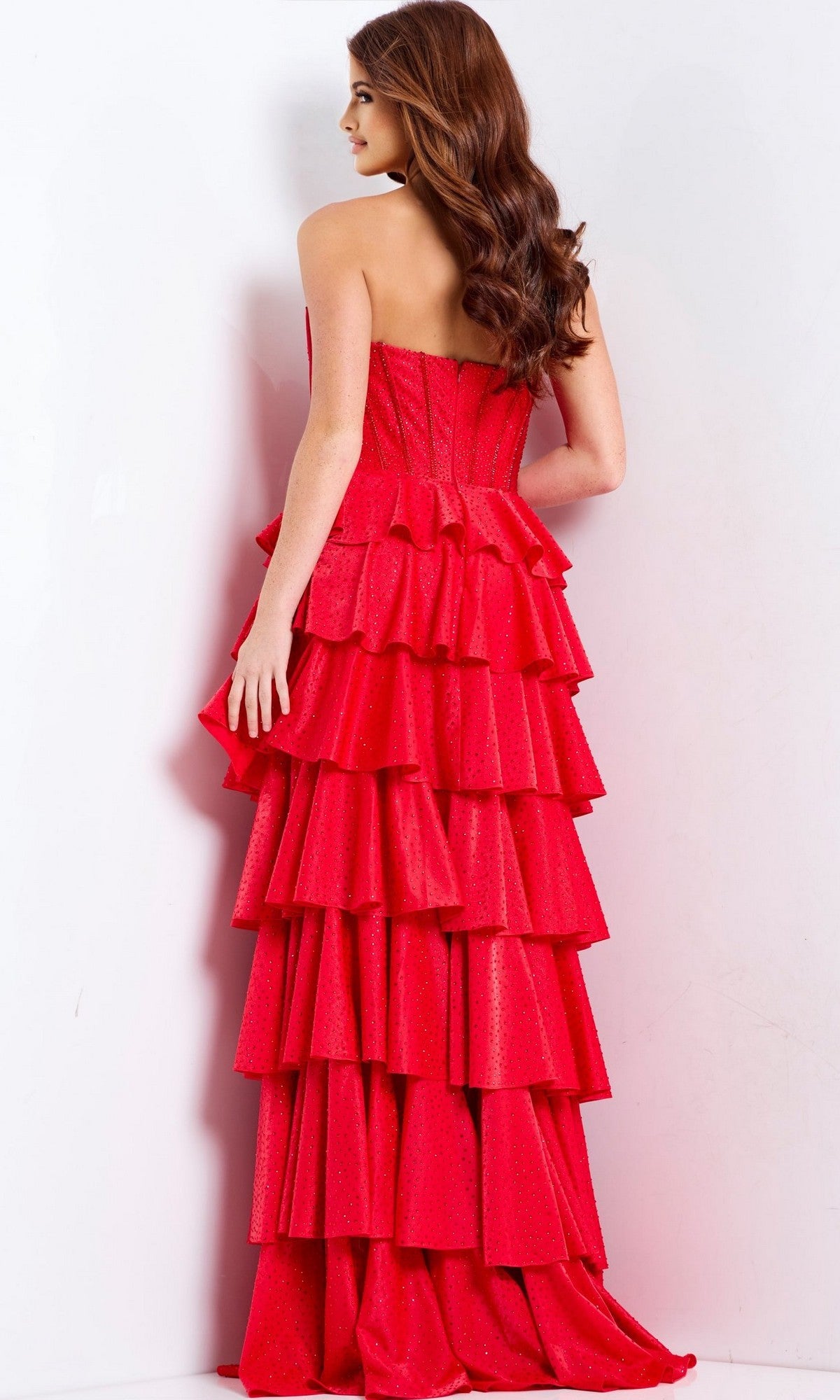 A woman with long brown hair stands facing away, wearing the Jovani 49780 long prom dress, which is strapless with a sweetheart neckline and features bold red, multi-tiered ruffles.